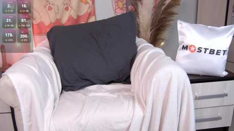 Media: Video of a cozy bedroom scene featuring a white armchair draped in a white blanket, a gray pillow, and a \"MOSTBET\" pillow on a white dresser with a potted plant in the background.