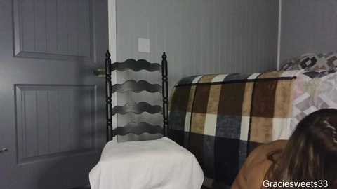 Media: A video of a rustic, grey-painted room with a metal-framed chair, white tablecloth, and a plaid-patterned blanket on a bed.