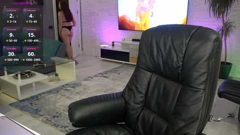 Media: Video of a modern living room with a black leather recliner, white brick wall, TV screen, and a woman in a bikini standing.