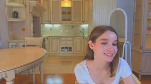 Media: Video of a young Caucasian woman with straight brown hair and a light complexion, smiling in a spacious, elegant kitchen with beige cabinets, wooden floor, and a round wooden table.