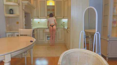 Media: A video of a modern, spacious kitchen with beige cabinets, a woman in a black thong standing at the counter, wooden floor, and a wicker chair.