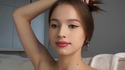 Media: Video of a young Asian woman with fair skin, brown hair, and light makeup, posing with her hand behind her head, wearing a white strapless top, in a modern, softly lit room.