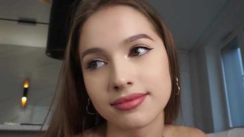 Media: Video of a young woman with light brown skin, straight brown hair, and brown eyes, wearing subtle makeup and silver hoop earrings, smiling gently in a modern, well-lit kitchen with stainless steel appliances.