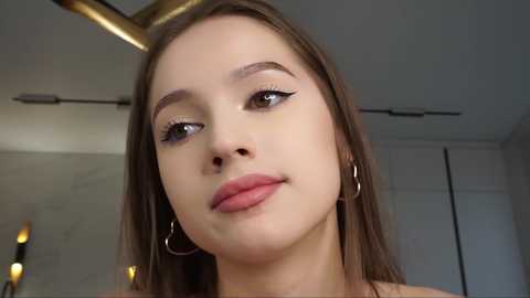 Media: Video of a young Caucasian woman with fair skin, long brown hair, wearing subtle makeup, hoop earrings, and a white top, indoors with a modern, minimalist background.