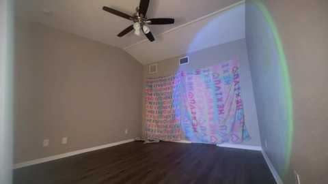 Media: Video of a sparse, empty room with beige walls, a dark hardwood floor, a ceiling fan, and a colorful, patterned curtain on the right wall.