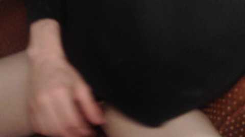 Media: Video of a close-up view of a person's hand resting on a thigh, wearing a black long-sleeve shirt. The background features a textured fabric with a reddish-brown pattern.