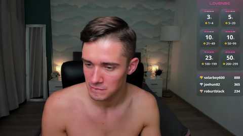 Media: Video of a shirtless, young, light-skinned man with short brown hair, sitting indoors. Background shows a dimly lit room with a bed, lamp, and digital readout of streaming stats.