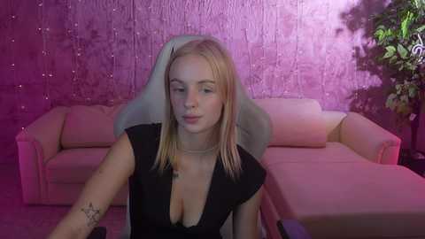 Media: Video of a blonde, fair-skinned woman with medium-sized breasts, wearing a low-cut black dress, sitting on a white chair with a star tattoo on her arm. She has a serene expression. The background features a pink textured wall and a beige couch.