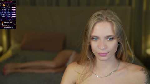 Media: Video of a fair-skinned, blonde woman with blue eyes, topless, wearing a necklace, standing in a dimly-lit bedroom with a bed and pillows in the background.