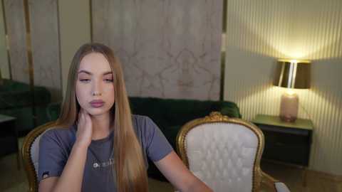 Media: Video of a young woman with long, straight brown hair, wearing a gray t-shirt, sitting at a table in a dimly-lit, elegant living room with a green couch, a gold-framed chair, and a lamp.