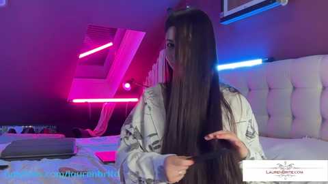 Media: Video of a woman with long, dark hair, wearing a light jacket, in a modern, neon-lit bedroom, adjusting her hair with a black hairbrush.