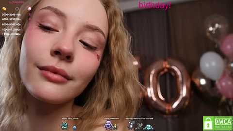 Media: Video of a young woman with fair skin, wavy blonde hair, and closed eyes, wearing pink eyeshadow. Background features a blurred, decorated room with metallic balloons.