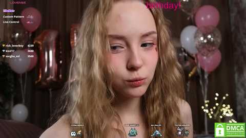 Media: Video of a young, fair-skinned, blonde girl with wavy hair, wearing a light-colored top, in a dimly lit room with pink and silver balloons.