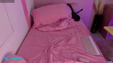 Media: Video of a pink-themed bedroom with a bed covered in pink bedding, a white keyboard on the floor, a black electric guitar leaning against the wall, and a yellow table in the background.