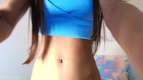 Media: A video of a young woman with long brown hair, wearing a blue crop top, showcasing her toned midriff and slight belly button, standing in a brightly lit room with pastel-colored wall art in the background.