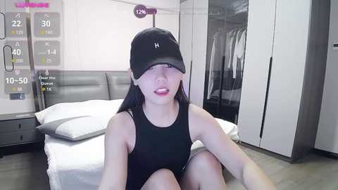 Media: Video of an Asian woman in a black tank top, sitting on a bed, wearing a black cap with a white \"H\" logo, in a modern bedroom with white furniture and a gray headboard.