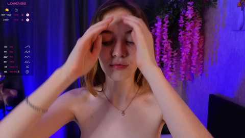 Media: Video of a young, fair-skinned woman with light brown hair, topless, adjusting her hair with her hands. Background features purple and pink floral arrangements, dimly lit room, and streaming platform interface.