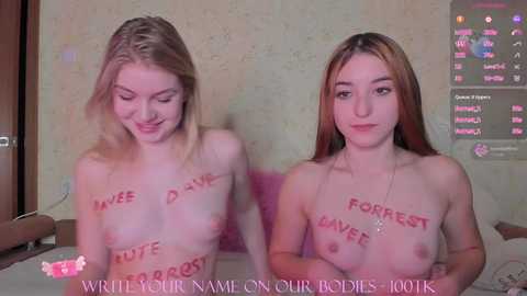 Media: Video of two young, fair-skinned, topless women with blonde hair, holding hands, with red \"F**k me\" tattoos on their chests. Background shows a pink bed and a TV screen displaying \"Forrest Gump\" on a beige wall.