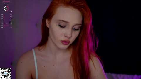 Media: Video of a fair-skinned, red-haired woman with long hair, wearing a white spaghetti-strap top, looking down with a contemplative expression. Background is dark, with purple lighting.