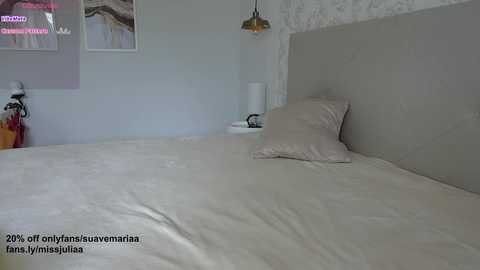 Media: A video of a minimalist bedroom with a large, neatly made bed covered in white linens. The room features a light grey upholstered headboard, white walls adorned with abstract art, and a small white nightstand with a lamp.