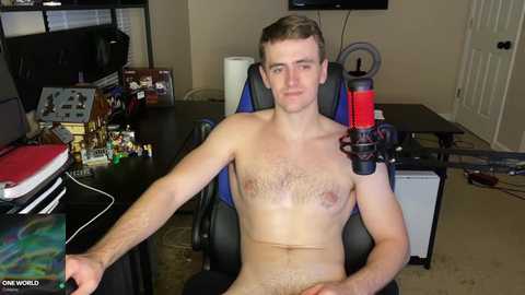 Media: Video of a shirtless, fair-skinned, muscular young man with short brown hair, seated in a blue gaming chair, holding a red microphone, surrounded by gaming equipment in a cluttered room.