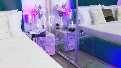 Media: Video of a modern bedroom with a white bed, clear acrylic chairs, white nightstands, purple and blue mood lighting, and floral decorations.