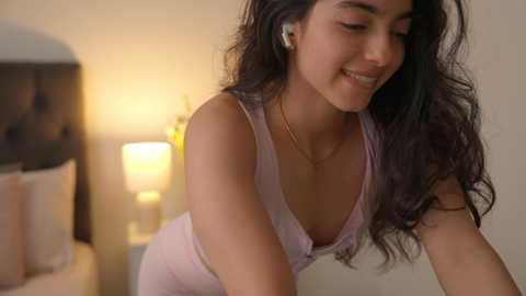 Media: Video of a young woman with long, wavy dark hair, wearing a pink sleeveless top, leaning forward, smiling, in a softly lit bedroom with a bed and nightstand.