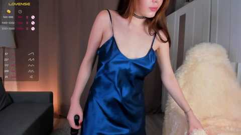 Media: Video of a fair-skinned, slender woman with long auburn hair wearing a blue satin slip dress, standing in a modern bedroom with a fluffy white rug and grey couch.