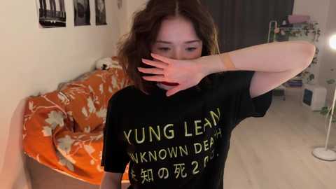 Media: A video of a young woman with shoulder-length brown hair, wearing a black t-shirt with yellow text, covering her face with one hand, standing in a dimly lit, cluttered room.