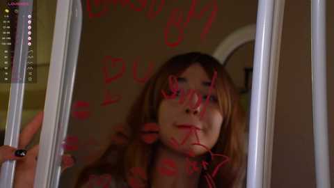 Media: Video of a young woman with light skin and long, brown hair, wearing a red lipstick. Red graffiti scribbled on a mirror reads \"Don't Be Stupid\" and \"Dumb.\