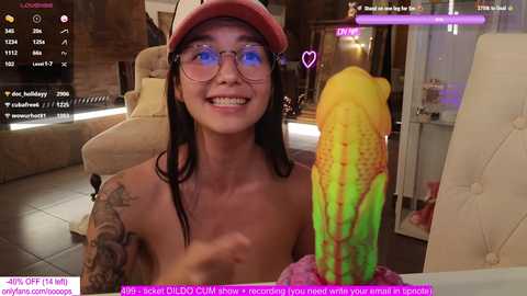 Media: Video of a topless, smiling Asian woman with glasses, wearing a pink baseball cap, holding a large, colorful dildo, in a modern living room with gaming elements on screen.