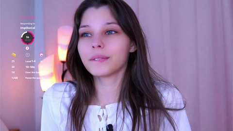 Media: Video of a young, fair-skinned woman with long brown hair, wearing a white blouse, standing indoors with a soft, pinkish background.