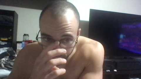 Media: Video of a bald, fair-skinned, glasses-wearing man covering his mouth, sitting shirtless in a dimly-lit room with a TV and cluttered shelves in the background.