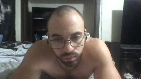 Media: Video of a bald, bearded, light-skinned man with glasses, lying shirtless on a bed, eyes closed, in a dimly lit bedroom with a TV and cluttered dresser.