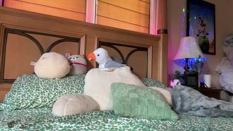 Media: Video of a cozy bedroom with a wooden headboard, a plush white pillow, a green blanket, a stuffed animal, and a parrot on a bed. The background includes a nightstand with a lamp and colorful decorations.