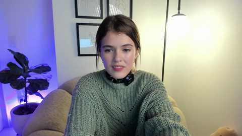 Media: Video of a young woman with light skin and brown hair, wearing a chunky knit sweater and a black choker, sitting on a beige couch. Background features a potted plant, framed art, and a glowing lamp.