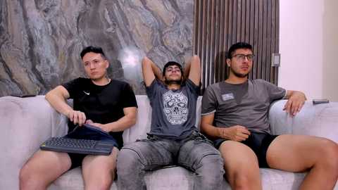 Media: Video of three men, two with short hair, one with glasses, sitting on a light couch, wearing casual attire, watching TV in a modern living room.