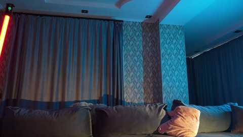 Media: Video of a dimly lit living room with a plush, gray sofa and a patterned wallpaper background, featuring a person sitting with a red light source casting a dramatic glow.