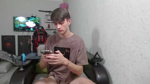 Media: Video of a young, fair-skinned man with short brown hair, wearing a brown T-shirt and shorts, sitting on a black couch in a dimly lit room. He's engrossed in his smartphone.