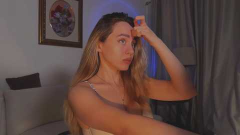 Media: A video of a young Caucasian woman with long, blonde hair, sitting in a dimly lit living room. She wears a beige top, adjusting her hair, with a framed painting and curtains in the background.