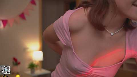 Media: Video of a woman with light skin and brown hair, wearing a pink, off-the-shoulder top, exposing her left breast. Background includes a lamp, pink bunting, and blurred furniture.