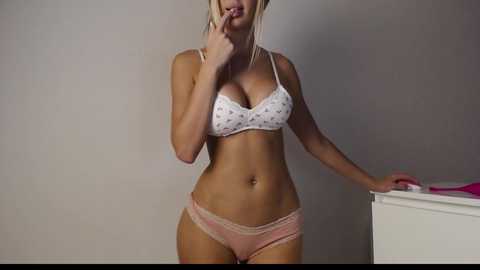 Media: Video of a blonde woman with fair skin, wearing a white bra with polka dots and pink lace panties, standing against a plain background. She's touching her lips suggestively.