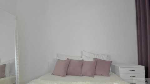 Media: A minimalist bedroom video featuring a white bed with a white duvet, three pink pillows, a white nightstand, and a mirror on the left wall.