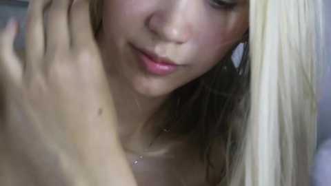 Media: Close-up video of a light-skinned woman with wet, blonde hair, gazing downwards with a soft smile. Her left hand gently touches her hair. Background is blurred, focusing on her face.