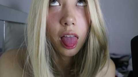 Media: Video of a blonde, fair-skinned woman with long, straight hair, sticking out her tongue. She has blue eyes and is topless, in a dimly lit bedroom with a bed and a mirror in the background.