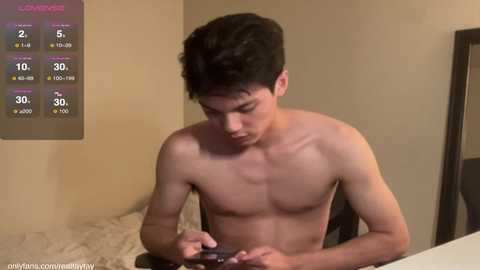 Media: Video of a shirtless young Asian man with short dark hair, focused on a phone, in a dimly lit room with beige walls and a weather forecast on a screen.