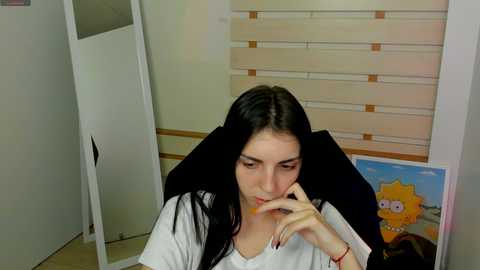 Media: Video of a young woman with long dark hair, wearing a white t-shirt, sitting with head on her hand, in an office with a poster of The Simpsons behind her.