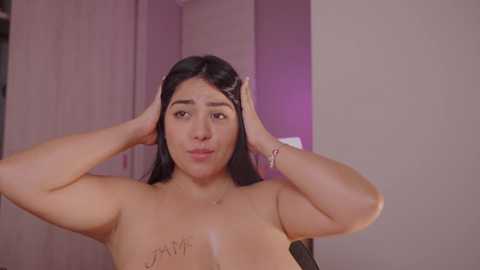 Media: Video of a topless Latina woman with medium brown skin, long black hair, and medium-sized breasts, adjusting her hair in a bedroom with a wooden wardrobe and purple lighting.