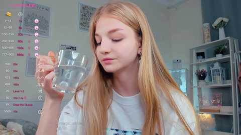 Media: Video of a young blonde girl in a white T-shirt, holding a glass of water, in a bright, minimalist room with a calendar and shelves.