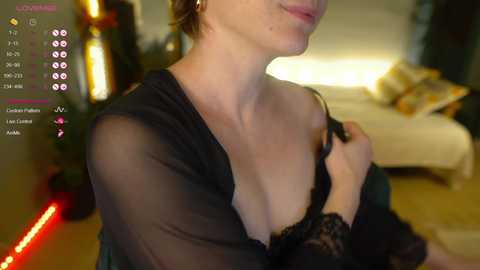 Media: A video of a fair-skinned woman with light brown hair, wearing a black sheer robe, taken indoors with a dimly lit room and a bed in the background.